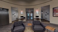 This single-story clubhouse is designed as a well-appointed community hub, combining functional amenities with a warm, inviting atmosphere. The interior is anchored by a distinctive curved rotunda-style layout, with smooth curved walls, a recessed ceiling detail, and neutral warm tones throughout that contribute to an open, cohesive feel.

The lounge area features comfortable upholstered armchairs arranged around a wood-accent coffee table, creating an ideal setting for relaxed socializing or quiet downtime. A wall-mounted flat-screen television is framed by a series of decorative built-in niches displaying decorative accents, adding visual interest and character to the space. Wood-style flooring runs throughout the interior, complementing the contemporary design palette.

A dedicated business center is integrated into the space, equipped with desktop computer workstations positioned along wall-mounted desks, illuminated by pendant and wall-mounted lighting. Decorative framed artwork adds a polished finish to the area.

Adjacent to the lounge, a kitchenette provides counter seating and cabinetry, offering a practical space for residents. Double glass-panel entry doors provide a clear sightline to the exterior.

Outside, a covered patio with rustic wood-framed doors leads directly to a generously sized outdoor pool and spa, surrounded by a spacious sun deck furnished with lounge chairs and umbrella-shaded seating, framed by mature trees and lush landscaping.