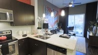 This single-floor space showcases a thoughtfully designed open-concept layout that seamlessly connects the kitchen and living areas. The kitchen is outfitted with rich dark cabinetry that contrasts sharply against light marble-veined countertops, creating a striking visual dynamic. A double stainless steel sink sits beneath stylish pendant lighting, adding both function and flair. Stainless steel appliances, including a dishwasher, further enhance the kitchen's modern appeal.

The kitchen flows naturally into the living area, where a ceiling fan with a sleek profile anchors the space above. A large, bold piece of artwork featuring vivid orange and grey tones serves as a dramatic focal point on the accent wall, adding an energetic, urban character to the interior. Dark walls contrast with lighter furnishings in the living area, contributing to a moody yet sophisticated ambiance.

A large window allows natural light to filter in, brightening the living space and offering a view of the outdoors. The overall design balances contemporary aesthetics with functional living, making this a visually cohesive and well-appointed space.