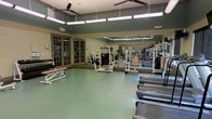 This fitness center offers a spacious, single-floor layout designed to accommodate a full range of exercise activities. The room is furnished with a comprehensive selection of strength-training machines and cardio equipment, including Precor treadmills, cable machines, and various resistance stations, providing users with everything needed for a complete workout routine.

Large mirrored walls line one side of the room, enhancing the sense of openness and allowing users to monitor their form during exercise. The flooring is a durable, cushioned green surface well-suited for gym use, offering both comfort and safety underfoot.

Overhead, ceiling fans and fluorescent lighting ensure the space remains well-lit and comfortably ventilated throughout. Elegant French-style glass doors along one wall lend a refined architectural touch, adding character to what might otherwise be a purely functional environment.

The overall ambiance is clean, organized, and inviting, with ample space between equipment to allow for comfortable movement and use by multiple individuals simultaneously. The thoughtful combination of practical fitness amenities and classic design details makes this fitness center both functional and visually appealing.