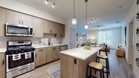 Located at 4560 Mission Gorge Pl in San Diego, CA, this single-story residence offers a thoughtfully designed layout with modern finishes throughout. The kitchen is a standout feature, showcasing warm wood-toned cabinetry with matte black hardware, white quartz countertops, a textured tile backsplash, and a full suite of stainless steel appliances including a gas range, over-the-range microwave, and refrigerator. A central island with pendant lighting provides additional prep space and casual seating, seamlessly connecting the kitchen to the open living and dining area. The living room is bright and welcoming, with large windows and a glass door that invite natural light and offer views of palm trees and a pool area beyond. Light wood-style flooring runs throughout the main living spaces, adding warmth and continuity. The bedrooms are carpeted for comfort and feature windows with outdoor greenery views, creating a calm, restful atmosphere. The bathroom includes a wood-tone vanity, soaking tub with shower, and clean tile finishes. A hallway laundry closet houses a stacked washer and dryer unit, adding everyday convenience. Overall, the space presents a clean, contemporary aesthetic well-suited to San Diego living.