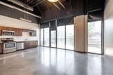 Located at 5472 Wild Rose Ln in West Des Moines, IA, this single-floor unit offers a distinctive industrial-modern aesthetic that blends open living space with functional design. The main living area features polished concrete floors and soaring exposed ceilings with visible ductwork and structural elements, creating an urban loft-like atmosphere. Large floor-to-ceiling windows and a glass door fill the interior with natural light while offering views of the surrounding cityscape. A pendant light fixture anchors the open main area, lending warmth to the industrial surroundings. The kitchen is well-appointed with rich dark wood cabinetry, stainless steel appliances including a refrigerator, range, microwave, and dishwasher, complemented by light countertops and a single-basin sink. A dedicated closet space features dual sections with wire shelving and hanging rods, providing organized storage. An outdoor patio accessed via a glass door offers an open-air retreat with horizontal wood railing details and views of the neighboring commercial district. The unit's brick exterior and modern finishes reflect the broader architectural character of the building, making this a compelling and functional urban living space.