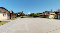 This residential community is designed with outdoor living and recreation in mind. At its center lies a generously sized concrete basketball court, complete with multiple hoops, offering residents a dedicated space for athletic activity and casual play. The court is flanked on both sides by the community's residential buildings, which feature a mix of one- and two-story structures finished in neutral earth tones with brown trim and pitched roofs, lending the complex a cohesive and approachable aesthetic.

Surrounding the court and buildings, mature trees provide natural shade and greenery, softening the hardscape and contributing to a welcoming atmosphere. Patches of lawn are interspersed throughout the common areas, enhancing the sense of an open, community-oriented environment. The layout encourages an active lifestyle while maintaining a sense of neighborly proximity between units.

The open sky and abundant natural light typical of a sunny Southern California-style climate bathe the entire complex throughout the day, creating a bright and inviting outdoor setting. Walkways connect the various sections of the community, making it easy to navigate between residential units and shared amenities. Overall, the property presents a functional and community-focused living environment well-suited for residents who value outdoor space and recreational access.
