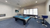This contemporary recreational lounge is designed to offer a well-rounded entertainment experience across a single, open floor. The space is anchored by a full-size pool table with pendant lighting overhead, complemented by a shuffleboard table positioned near large windows that invite ample natural light. Blue patterned carpet tiles run throughout, tying together the cohesive, modern aesthetic.

A dedicated lounge area features a plush blue sofa, colorful modular ottomans, and a wall-mounted flat-screen TV with a soundbar, making it ideal for casual viewing and relaxation. Adjacent to the lounge, a striking white stacked-stone accent wall frames a sleek linear fireplace, adding warmth and texture to the space.

A bar-height counter with bold blue swivel stools serves as a social hub, while a wall-mounted TV and vibrant abstract artwork contribute to the energetic atmosphere. A foosball table offers additional gaming options, and a separate glass-walled room visible through the entrance provides a quieter, semi-private area for focused activities.

Recessed lighting throughout maintains a bright, welcoming ambiance, and the open layout allows for fluid movement between activity zones. The overall design balances modern aesthetics with functional, community-oriented amenities.