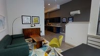 This single-floor space showcases an open-concept layout that seamlessly connects the living area and kitchen, creating a cohesive and functional environment. The living area features soaring ceilings that amplify the sense of space, complemented by crisp white walls contrasted against a deep charcoal accent wall. A geometric-patterned area rug in teal, yellow, and grey anchors the seating area, which includes a plush sofa and a striking chartreuse accent chair that adds a bold pop of color. A solid wood coffee table grounds the space with warmth. Framed artwork decorates the walls, contributing to the curated aesthetic. The kitchen is outfitted with dark cabinetry, stainless steel appliances, and a tiled backsplash, with a light-toned countertop that provides contrast. A wall-mounted mini-split unit ensures climate control. The flooring throughout is light-toned hardwood, lending continuity between the living and kitchen zones. Recessed lighting provides consistent illumination, while the overall design balances modern minimalism with carefully selected furnishings for a polished, contemporary ambiance.