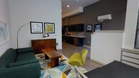 This single-floor space showcases an open-concept layout that seamlessly connects the living area and kitchen, creating a cohesive and functional environment. The living area features soaring ceilings that amplify the sense of space, complemented by crisp white walls contrasted against a deep charcoal accent wall. A geometric-patterned area rug in teal, yellow, and grey anchors the seating area, which includes a plush sofa and a striking chartreuse accent chair that adds a bold pop of color. A solid wood coffee table grounds the space with warmth. Framed artwork decorates the walls, contributing to the curated aesthetic. The kitchen is outfitted with dark cabinetry, stainless steel appliances, and a tiled backsplash, with a light-toned countertop that provides contrast. A wall-mounted mini-split unit ensures climate control. The flooring throughout is light-toned hardwood, lending continuity between the living and kitchen zones. Recessed lighting provides consistent illumination, while the overall design balances modern minimalism with carefully selected furnishings for a polished, contemporary ambiance.