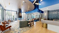 Located at 1201 Callowhill St in Philadelphia's Callowhill neighborhood, this expansive single-floor amenity space is a striking blend of contemporary design and vibrant personality. The interior is defined by bold cobalt blue accent walls and ceilings that contrast beautifully with warm light oak hardwood flooring and multicolored terrazzo-patterned carpeting throughout the lounge areas. A dramatic curved ceiling detail anchors the central lounge, where a freestanding pendant fireplace serves as a stunning focal point surrounded by an eclectic mix of seating—including cognac leather chairs, rich red velvet sofas, and neutral accent chairs—creating an inviting, residential-inspired atmosphere. Large street-facing windows flood the space with natural light and offer views of the surrounding streetscape. A dedicated kitchenette area features a white island with mosaic tile detailing, wood-paneled cabinetry, and a suite of modern pendant lighting with gold-toned fixtures. A vibrant abstract mural adds artistic energy to the co-working lounge zone, furnished with navy velvet chairs and warm wood tables. The glass-enclosed conference room provides a private meeting space with a solid wood table, navy upholstered seating, and sophisticated globe pendant lighting. A staffed reception desk with dark mosaic tile backing completes this thoughtfully designed, amenity-rich space.