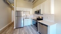 Located on Smallman Street in Pittsburgh's vibrant Strip District, this single-floor unit masterfully combines industrial heritage with contemporary design. The space is defined by its exposed painted brick walls, which lend a raw, textured character throughout, while light wood-tone flooring provides a warm contrast underfoot.

The kitchen is a standout feature, outfitted with a full suite of stainless steel appliances, including a refrigerator, electric range, microwave, and dishwasher. White shaker-style cabinetry reaches toward the ceiling, maximizing storage, while a classic white subway tile backsplash and light quartz countertops complete the clean, modern aesthetic. The layout makes efficient use of space with a peninsula-style counter section.

The living area benefits from natural light streaming through a roller shade-equipped window that frames views of the surrounding neighborhood's historic architecture. The exposed brick wall in this room adds to the industrial loft character of the unit.

The bathroom is neatly appointed with a white vanity, large mirror with overhead lighting, and light tile flooring, maintaining a cohesive, clean palette throughout. Overall, the space offers a refined urban living experience rooted in Pittsburgh's industrial past.
