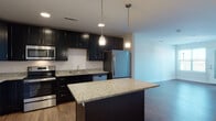 Located at 26 Kettle Point Ave in East Providence, Rhode Island, this single-floor unit offers a thoughtfully designed open-concept layout with modern finishes throughout. The living area is anchored by rich hardwood-style flooring that extends across the main living and dining spaces, creating a warm and cohesive aesthetic. Large windows and a glass-paneled entry door allow natural light to fill the room, with views extending to an outdoor balcony area.

The kitchen is a standout feature, outfitted with dark espresso cabinetry that reaches toward the ceiling, providing generous storage. Speckled granite countertops run throughout, complemented by a central island with matching surfaces and pendant lighting overhead. A full suite of stainless steel appliances—including a refrigerator, range, built-in microwave, and dishwasher—completes the functional and stylish cooking space.

The bedroom is carpeted for comfort and features a window with balcony views, offering a quiet retreat. The bathroom is finished with classic black-and-white hexagonal mosaic tile flooring, a clean white vanity with dark cabinetry, and a full tub-shower combination. A dedicated laundry closet houses a stacked full-size washer and dryer, adding everyday convenience to this well-appointed home.