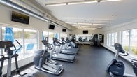 This modern fitness center is designed to provide a comprehensive workout environment in a clean, well-organized single-floor layout. The space is lined with large windows along one wall, allowing natural light to fill the room and offering views of an outdoor pool area, creating an open and energizing atmosphere.

The cardio zone is prominently featured, housing a row of treadmills and elliptical machines from leading fitness brands, all neatly arranged for easy access. Overhead, bright fluorescent strip lighting ensures excellent visibility throughout the facility, complemented by an exposed industrial-style ventilation duct that adds a contemporary aesthetic touch.

A full wall of mirrors on the far end of the space visually expands the room while allowing users to monitor their form during workouts. Multiple flat-screen TVs are mounted at strategic points throughout the facility, providing entertainment during exercise sessions.

The strength training section is situated toward the rear, equipped with a multi-station cable and weight machine system suitable for a full-body workout. Dark rubberized flooring covers the entire space, offering both durability and safety. The overall ambiance is bright, functional, and welcoming, making this fitness center a well-rounded facility for a wide range of exercise needs.