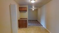 Derby Estates is a single-floor residence that combines straightforward design with functional living. The space transitions naturally from the kitchen area into the living space, creating an open, connected feel throughout. The kitchen features warm-toned wooden cabinetry, including both upper and lower cabinet storage with a countertop workspace, providing ample room for everyday use. The flooring in the kitchen area consists of light tile, offering durability and easy maintenance. Moving into the living area, the flooring transitions to carpet, adding a layer of comfort underfoot. A ceiling fan is centrally positioned in the living space, ensuring comfortable airflow year-round. The walls are finished in a soft, neutral tone that complements the cabinetry and flooring throughout, lending a cohesive and calm ambiance to the interior. The textured ceiling adds subtle visual interest to the space. Overall, Derby Estates presents a practical and neatly arranged single-floor layout suited for comfortable day-to-day living, with thoughtful details that make the most of its compact footprint.