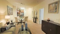 This single-floor residence offers an open-concept layout that integrates the living, dining, and kitchen areas into one cohesive and functional space. The interior is finished in warm, neutral tones, with beige carpeting throughout the main living areas that contributes to a welcoming ambiance.

The living area is furnished with comfortable seating and a low-profile coffee table, anchored by a dark media console that provides both storage and display space. Adjacent to this is a dedicated dining area featuring a glass-top table surrounded by elegant high-back upholstered chairs, ideal for both casual and formal dining.

The kitchen is visible from the dining space and is equipped with stainless steel appliances, including a refrigerator, and features warm wood cabinetry. A peninsula with pendant lighting above serves as a natural transition between the kitchen and dining areas.

A central entryway corridor leads toward what appear to be additional rooms, flanked by a decorative console table topped with a sunburst mirror. Track lighting and flush-mount ceiling fixtures provide ample illumination throughout, enhancing the bright, airy feel of the space.