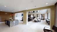 The Social Lounge is a thoughtfully designed, single-floor communal space that blends contemporary style with comfortable functionality. The open-plan layout creates a seamless flow between distinct zones, allowing residents and guests to move freely throughout the space.

Upon entering, a striking granite-topped bar counter anchors one side of the room, its richly veined stone surface adding a touch of elegance. Pendant lighting in warm amber tones hangs above, creating an inviting atmosphere. Adjacent to the bar, a lounge area features a wall-mounted flat-screen television set against a textured tile accent wall, providing a relaxed entertainment zone.

The dining area is furnished with a long, dark-framed table surrounded by upholstered white chairs, offering ample seating for group gatherings. A second flat-screen television is visible in this zone, enhancing its versatility. Natural light filters in through multiple windows, brightening the neutral color palette of warm beige tones and light-colored tile flooring throughout.

Structural columns define zones without enclosing the space, maintaining an airy, open feel. The overall ambiance is modern, clean, and welcoming — well-suited for social events and everyday community interaction.