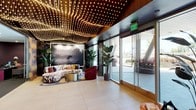 This sophisticated lobby makes an immediate impression with its extraordinary ceiling installation — thousands of warm pinpoint lights arranged in sweeping, wave-like patterns across a dark field, evoking a luminous night sky. The space is anchored by large-format floor tiles in a warm neutral tone, providing a clean, polished foundation that lets the bold design elements shine.

A central lounge area features a curved, vibrantly patterned sofa paired with a deep burgundy accent chair and a brushed gold cylindrical coffee table, all positioned beneath a dramatic large-scale black-and-white photographic artwork. Lush tropical plants in substantial planters are thoughtfully distributed throughout, adding organic warmth and texture.

A secondary seating nook offers a more intimate atmosphere, set against a rich plum-colored accent wall with a stainless steel elevator as a focal point. Here, sleek leather-trimmed chairs and a glass-top table rest on a jewel-toned area rug, accompanied by a large digital display wall. Iridescent curtain panels serve as elegant space dividers, catching light with a prismatic shimmer.

Floor-to-ceiling glass walls and automatic entry doors flood the space with natural daylight and provide unobstructed views of the exterior surroundings, seamlessly connecting indoors and outdoors. The overall ambiance is energetic yet refined — a curated blend of contemporary art, luxurious materials, and dynamic lighting design.