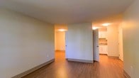 This single-floor residence presents a clean, functional layout designed for comfortable everyday living. The main living area is finished with warm-toned hardwood flooring that extends throughout, creating a cohesive and inviting atmosphere. Neutral-painted walls and flush ceiling lighting contribute to a bright, well-illuminated interior.

The open layout connects the primary living space to an adjacent room, offering flexibility in how the space can be arranged and utilized. A central dividing wall adds a sense of architectural definition while maintaining an airy, open feel throughout.

The kitchen is accessible through a nearby doorway and features white cabinetry, a sink, and light countertops, providing a practical and tidy cooking space. The cabinetry offers ample storage, and the layout is straightforward and efficient.

Doorways throughout the unit are framed in white trim that complements the overall clean aesthetic. The consistent use of neutral tones, hardwood-style flooring, and recessed ceiling fixtures gives the entire space a cohesive, move-in-ready appearance. Overall, this residence offers a simple yet well-appointed interior suited to a variety of living needs.