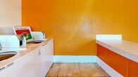 Located at 2433 W Campbell Ave in Phoenix, Arizona, this single-floor laundromat offers a compact yet efficiently organized space designed for self-service laundry needs. The interior is defined by its bold, warm aesthetic — a striking orange ceiling contrasts with crisp white walls, creating a vibrant and easily recognizable environment. Light tile flooring runs throughout, contributing to a clean and practical finish underfoot.

The main area is anchored by a row of front-loading washing machines along one wall, complemented by a set of stacked dryer units in an adjacent section, maximizing the use of available floor space. A centralized payment kiosk provides convenient card or coin-based access to the machines. Instructional and informational signage is prominently displayed on the walls, helping customers navigate machine usage and laundry best practices.

A tiled service counter with warm orange wood-panel accents runs along one side of the room, offering a dedicated folding and sorting surface. Natural light enters through several windows fitted with security bars, keeping the space well-lit while maintaining safety. A security door at the entrance features a decorative sunburst grille design, adding a touch of character to the utilitarian space. The layout is open and straightforward, ensuring ease of movement and a functional customer experience.