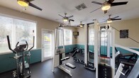 This fitness center is a dedicated, single-floor exercise facility designed to support a comprehensive workout routine. The room features a distinctive two-tone wall treatment, with a rich teal accent along the lower half and a warm neutral tone above, giving the space a clean and energizing aesthetic. Dark rubber flooring covers the entire area, providing both durability and safety underfoot.

Natural light fills the room through multiple large windows fitted with horizontal blinds, offering views of the surrounding outdoor greenery. Several ceiling fans are strategically distributed throughout the space, ensuring consistent airflow and comfort during workouts.

The facility is outfitted with an impressive array of equipment. Cybex-brand strength machines, including cable and weight stack units, occupy a central position in the room, while a multi-functional cable crossover station serves as a focal point. Additional strength equipment includes adjustable benches and a seated press machine. On the cardio side, the space features a treadmill and an elliptical trainer, providing options for both low- and high-impact aerobic exercise.

A wall-mounted television is positioned for visibility across the room, and posted signage provides facility guidelines for users. With direct door access to the outdoors and ample equipment for both strength training and cardiovascular fitness, this facility offers a practical and well-organized environment for exercise.