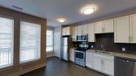 This single-floor space offers a thoughtfully designed interior with a strong emphasis on modern finishes and functional layout. The kitchen is a standout feature, showcasing crisp white shaker-style cabinetry that extends to the ceiling, providing generous storage throughout. Stainless steel appliances — including a refrigerator, electric range, and over-the-range microwave — lend a sleek, contemporary feel. A dark slate mosaic tile backsplash adds texture and contrast behind the range, while granite countertops and a stainless steel sink complete the polished look. Dark hardwood flooring runs throughout, anchoring the space with warmth and depth. Flush-mount ceiling light fixtures provide consistent overhead illumination, complementing the natural light that filters in through large windows dressed with horizontal blinds. The neutral wall tones in warm taupe and crisp white create a balanced, inviting ambiance. A paneled interior door is visible, hinting at a well-organized floor plan. Overall, this space combines practical design with quality materials, making it a comfortable and visually cohesive environment suited for everyday living.