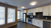 This single-floor space offers a thoughtfully designed interior with a strong emphasis on modern finishes and functional layout. The kitchen is a standout feature, showcasing crisp white shaker-style cabinetry that extends to the ceiling, providing generous storage throughout. Stainless steel appliances — including a refrigerator, electric range, and over-the-range microwave — lend a sleek, contemporary feel. A dark slate mosaic tile backsplash adds texture and contrast behind the range, while granite countertops and a stainless steel sink complete the polished look. Dark hardwood flooring runs throughout, anchoring the space with warmth and depth. Flush-mount ceiling light fixtures provide consistent overhead illumination, complementing the natural light that filters in through large windows dressed with horizontal blinds. The neutral wall tones in warm taupe and crisp white create a balanced, inviting ambiance. A paneled interior door is visible, hinting at a well-organized floor plan. Overall, this space combines practical design with quality materials, making it a comfortable and visually cohesive environment suited for everyday living.