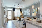 This thoughtfully designed single-floor residence offers a seamless blend of modern finishes and functional living spaces. The open-concept layout connects the kitchen and living area, creating a bright, cohesive environment ideal for both everyday living and entertaining. The kitchen is outfitted with crisp white cabinetry, granite countertops, a subway tile backsplash, and a full suite of stainless steel appliances, including a range, over-the-range microwave, dishwasher, and refrigerator. A kitchen island with pendant lighting serves as both a workspace and casual dining area. Wood-look flooring flows throughout the main living spaces, while crown molding and ceiling fans add refined architectural detail. The living room is bathed in natural light from large windows dressed with horizontal blinds and navy drapes, with a sliding glass door providing access to a private balcony. The bedroom features warm, bold accent walls, plush carpeting, a ceiling fan, and generous natural light. A spacious walk-in closet offers ample storage with multi-tiered hanging rods. The en-suite bathroom is finished with a dual-sink granite vanity, white cabinetry, and wood-look flooring, providing a clean and functional retreat directly accessible from the bedroom.