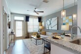 This thoughtfully designed single-floor residence offers a seamless blend of modern finishes and functional living spaces. The open-concept layout connects the kitchen and living area, creating a bright, cohesive environment ideal for both everyday living and entertaining. The kitchen is outfitted with crisp white cabinetry, granite countertops, a subway tile backsplash, and a full suite of stainless steel appliances, including a range, over-the-range microwave, dishwasher, and refrigerator. A kitchen island with pendant lighting serves as both a workspace and casual dining area. Wood-look flooring flows throughout the main living spaces, while crown molding and ceiling fans add refined architectural detail. The living room is bathed in natural light from large windows dressed with horizontal blinds and navy drapes, with a sliding glass door providing access to a private balcony. The bedroom features warm, bold accent walls, plush carpeting, a ceiling fan, and generous natural light. A spacious walk-in closet offers ample storage with multi-tiered hanging rods. The en-suite bathroom is finished with a dual-sink granite vanity, white cabinetry, and wood-look flooring, providing a clean and functional retreat directly accessible from the bedroom.