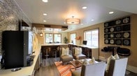 This single-floor amenity space combines a welcoming lounge area with a functional business center in a cohesive, well-appointed design. The interior is finished with rich dark hardwood flooring that contrasts elegantly against warm beige walls, creating an inviting and polished atmosphere throughout.

The lounge area is furnished with upholstered seating in neutral tones, accented by patterned throw pillows and a vibrant orange-striped area rug that adds a pop of color and visual warmth. A central coffee table anchors the seating arrangement, making the space ideal for casual gatherings or relaxed conversation.

Adjacent to the lounge, a built-in business center features a long countertop workstation lined with computer monitors and high-back bar-style seating. This area is bathed in natural light from a row of windows that line the wall, offering views to the exterior. A second counter space nearby, framed by hanging pendant lights in a darker finish, suggests an additional workspace or service area with bar-height seating.

Overhead, a combination of recessed lighting and a prominent drum pendant fixture provide warm, layered illumination throughout the space. Decorative elements — including tall ceramic vases, potted greenery, and wall-mounted ornamental pieces — complete the refined yet comfortable aesthetic of this thoughtfully designed communal environment.