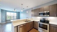 This single-floor unit is designed with a clean, contemporary aesthetic highlighted by soft blue-gray walls, wood-style flooring, and an open layout that promotes a sense of connectivity between spaces. The kitchen serves as a central feature, equipped with a full suite of stainless steel appliances including a refrigerator, electric range, built-in microwave, and dishwasher. Dark wood-grain cabinetry with sleek bar-pull hardware pairs with light quartz countertops to create a modern contrast. A peninsula with an extended countertop separates the kitchen from the living area, offering additional workspace and a natural gathering point. Pendant lights hang above the peninsula, adding focused illumination and a design-forward touch. A ceiling fan in the living area aids in air circulation, while a large window invites natural light and views of the surrounding neighborhood. The bedroom is a well-proportioned, quiet space with wood-style flooring, a ceiling fan, and a large window providing ample daylight. The bathroom is cleanly finished with tile flooring, a wood-grain vanity with a white countertop, a large mirror, and a shower-tub combination. The overall ambiance is modern, functional, and well-appointed.