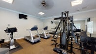 Located at 202 Stonemill Dr in Lynchburg, Virginia, this single-floor fitness center is thoughtfully designed to support a comprehensive workout experience. The space is finished with durable carpet tile flooring throughout and features crown molding along the ceiling line, lending a polished look to the functional environment. Neutral gray walls and flush-mounted lighting fixtures keep the room bright and clean in appearance.

The fitness center is divided into distinct workout zones. The cardio area houses multiple Precor treadmills positioned in front of a large wall mirror, which visually expands the space and allows users to monitor their form. An Octane elliptical trainer and a True upright stationary bike provide additional low-impact cardio options. A multi-station cable strength machine anchors the strength training section, complemented by free weights, kettlebells, and a stability ball.

Large windows along one wall admit generous natural light and offer views of an outdoor pool area. A ceiling fan overhead aids in air circulation during workouts. Supplementary amenities include a water cooler, an open-cubby storage unit stocked with small fitness accessories, and a wall-mounted television. The room also features a dedicated entry door, making the space feel self-contained and purposeful.