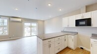 Located in Scarborough, Maine, this thoughtfully designed single-floor unit offers a bright and efficient layout comprising a living room, kitchen, bathroom, and closet. The open-concept design seamlessly connects the kitchen and living area, creating a cohesive and functional living space.

The kitchen is a standout feature, showcasing white shaker-style cabinetry with brushed gold hardware, granite countertops, a peninsula with additional seating and prep space, stainless steel appliances including a dishwasher, a built-in microwave, and a sink. Light hardwood flooring runs throughout the main living areas, while the entryway and bathroom feature elegant tile flooring.

The living room is bathed in natural light, courtesy of large windows and a sliding glass door that opens to views of the surrounding Scarborough neighborhood. A wall-mounted mini-split unit provides efficient climate control. The neutral warm-toned walls and recessed lighting throughout lend the space a clean, modern aesthetic.

The unit also includes a dedicated closet for storage and convenient access to additional spaces via the hallway. The overall design is fresh and move-in ready, reflecting quality new construction craftsmanship ideal for comfortable everyday living.