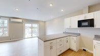 Located in Scarborough, Maine, this thoughtfully designed single-floor unit offers a bright and efficient layout comprising a living room, kitchen, bathroom, and closet. The open-concept design seamlessly connects the kitchen and living area, creating a cohesive and functional living space.

The kitchen is a standout feature, showcasing white shaker-style cabinetry with brushed gold hardware, granite countertops, a peninsula with additional seating and prep space, stainless steel appliances including a dishwasher, a built-in microwave, and a sink. Light hardwood flooring runs throughout the main living areas, while the entryway and bathroom feature elegant tile flooring.

The living room is bathed in natural light, courtesy of large windows and a sliding glass door that opens to views of the surrounding Scarborough neighborhood. A wall-mounted mini-split unit provides efficient climate control. The neutral warm-toned walls and recessed lighting throughout lend the space a clean, modern aesthetic.

The unit also includes a dedicated closet for storage and convenient access to additional spaces via the hallway. The overall design is fresh and move-in ready, reflecting quality new construction craftsmanship ideal for comfortable everyday living.