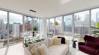 This elegantly designed high-rise residence showcases panoramic city skyline views through expansive floor-to-ceiling windows that flood the interior with natural light. The open-concept living and dining area is a standout, anchored by a plush white sectional sofa, a statement gold arc floor lamp, and a glass-top dining table with gold accents — all framed by unobstructed views of the urban landscape. The living space transitions seamlessly, with warm hardwood flooring and a wall-mounted entertainment unit complemented by curated artwork.

The kitchen is thoughtfully appointed with rich wood cabinetry, a large stone-topped island with built-in open shelving, and bar seating — ideal for casual dining. Stainless steel appliances and a subway tile backsplash complete the refined yet functional design.

The primary bedroom offers city views through generous windows, with light hardwood floors and a calm, neutral palette. A second bedroom features soft blush-toned walls and its own large window overlooking neighboring high-rises.

Both bathrooms are finished with terrazzo-style tile flooring, warm wood vanities with open shelving, and white countertops. One includes a glass-enclosed walk-in shower, while the other features a full soaking tub — delivering a spa-like atmosphere throughout.