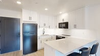 This efficiently designed single-floor unit offers a thoughtfully arranged layout encompassing an open-concept kitchen and living room, a hallway, and a full bathroom. The kitchen is a standout feature, showcasing crisp white shaker-style cabinetry extending to the ceiling, white quartz countertops, and a suite of black stainless steel appliances including a refrigerator, electric range, and over-the-range microwave. A central island with seating provides additional prep space and casual dining. Light wood-look flooring runs throughout, complementing the bright, white palette. The living area flows naturally from the kitchen and features a built-in entertainment unit with matching white cabinetry, a wall-mounted TV, and a wall-unit air conditioner. A window admits natural light and provides an outdoor view. The bathroom continues the modern design language with a white shaker vanity, quartz countertop, a large mirror with bar lighting, and a walk-in shower featuring marble-look tile walls and hexagonal mosaic floor tile with matte black fixtures. Recessed lighting throughout the unit enhances the clean, contemporary ambiance, making this a well-appointed and cohesive living space.