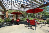 This outdoor amenity space is designed to offer residents a comprehensive leisure environment within a lushly landscaped, tree-lined setting. At the heart of the area is a resort-style swimming pool, surrounded by a spacious concrete deck furnished with lounge chairs and colorful red seating, ideal for relaxing in the sun. A prominent wooden pergola provides shaded gathering space beneath its slatted roof, where a round patio table surrounded by vibrant red chairs serves as a comfortable outdoor dining and socializing spot. A bold red umbrella adds a pop of color and additional shade to the seating area. The grounds are thoughtfully landscaped with manicured shrubs, seasonal plantings, and mature trees that create a natural, park-like atmosphere throughout. A dedicated grilling station with a charcoal grill and adjacent seating offers a convenient outdoor cooking area, perfect for casual gatherings. Beyond the pool, a fenced tennis court provides additional recreational options for active residents. The multi-story brick residential buildings surrounding the courtyard feature balconies that overlook the amenity area, reinforcing the community-centered design. Paved walkways connect the various outdoor areas, ensuring easy navigation throughout the grounds. The overall ambiance is bright, welcoming, and recreational, offering a variety of outdoor activities within a cohesive and well-maintained community setting.