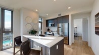 This contemporary single-floor residence showcases a thoughtfully designed open layout with high-end finishes throughout. The kitchen is a standout feature, equipped with dark navy cabinetry extending to the ceiling, stainless steel appliances including a built-in oven, microwave, and French door refrigerator, and a generous island with a crisp white quartz countertop and an integrated sink. Light wood-tone flooring runs throughout, complementing the clean, neutral palette. A floor-to-ceiling glass door in the kitchen connects the space to the outdoors, flooding the area with natural light and offering expansive city views.

The living area is anchored by dramatic floor-to-ceiling windows on two sides, framing sweeping urban and hillside panoramas. The open and airy atmosphere is enhanced by recessed lighting and smooth white walls throughout.

The bedroom continues the modern aesthetic, featuring large sliding glass doors that lead to a private balcony, bringing in abundant natural light and outdoor views. The bathrooms are finished with clean, minimalist design — one features a teal-blue floating vanity with a backlit mirror, while the other offers a white quartz countertop, an undermount sink, and a tiled walk-in shower, both reflecting a high standard of contemporary craftsmanship.
