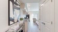 This single-floor studio unit presents a thoughtfully designed open-plan layout that maximizes every inch of available space. Light wood-look flooring flows continuously throughout, reinforcing a clean, cohesive aesthetic. The interior is bathed in natural light, largely thanks to a full-height sliding glass door fitted with vertical blinds that opens the living area to an outdoor view.

The galley-style kitchen runs along one wall and is outfitted with light wood cabinetry, granite-style countertops, a stainless steel sink, and stainless steel appliances including a refrigerator, microwave, and range. A glass console table near the entry adds a practical yet decorative touch, complemented by bold abstract wall art.

The living and sleeping areas share the open main space. A Murphy bed system integrates seamlessly with built-in storage, while a freestanding white wardrobe and a dedicated desk nook with wall-mounted television provide functional versatility. The arrangement allows the space to serve equally well as a living room or sleeping quarters.

The bathroom is finished in the same light, neutral palette, featuring a single-sink vanity with wood cabinetry and stone-look countertop, a walk-in shower with a curtain rod, and recessed lighting overhead. White walls and consistent flooring keep the space feeling open and airy throughout.