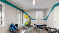 Located at 130 N Ridge Dr in Woodfin, NC, this dedicated exercise room is a well-equipped and thoughtfully designed fitness space. The room's interior is immediately distinguished by its bold teal accent stripe, which runs diagonally across the light gray walls, adding a dynamic and energetic visual character. Crown molding and a textured ceiling provide a polished finish, while flush linear lighting ensures the space is well-lit throughout.

The flooring features a stylish herringbone-patterned carpet tile in a mix of gray and teal tones, contributing both visual interest and a practical, durable surface for workouts. Natural light enters through multiple windows fitted with blinds, offering views of the surrounding neighborhood.

Equipment includes a Star Trac treadmill, a spin bike, a full rack of CAP hex dumbbells, a weight bench, a medicine ball stand with assorted balls, and resistance bands — providing a comprehensive range of cardio and strength training options. A glass-panel door connects the space to an adjacent room, enhancing flow and openness. Additional storage is available via a wicker basket. The overall ambiance is clean, vibrant, and functional, making this fitness room a practical and motivating environment for daily exercise.