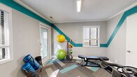 Located at 130 N Ridge Dr in Woodfin, NC, this dedicated exercise room is a well-equipped and thoughtfully designed fitness space. The room's interior is immediately distinguished by its bold teal accent stripe, which runs diagonally across the light gray walls, adding a dynamic and energetic visual character. Crown molding and a textured ceiling provide a polished finish, while flush linear lighting ensures the space is well-lit throughout.

The flooring features a stylish herringbone-patterned carpet tile in a mix of gray and teal tones, contributing both visual interest and a practical, durable surface for workouts. Natural light enters through multiple windows fitted with blinds, offering views of the surrounding neighborhood.

Equipment includes a Star Trac treadmill, a spin bike, a full rack of CAP hex dumbbells, a weight bench, a medicine ball stand with assorted balls, and resistance bands — providing a comprehensive range of cardio and strength training options. A glass-panel door connects the space to an adjacent room, enhancing flow and openness. Additional storage is available via a wicker basket. The overall ambiance is clean, vibrant, and functional, making this fitness room a practical and motivating environment for daily exercise.