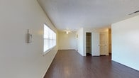 This single-floor space offers a clean, functional layout designed to maximize comfort and usability. The main living area is highlighted by rich, dark hardwood-style flooring that extends throughout, creating a warm and cohesive aesthetic. Cream-toned walls pair with a classic textured ceiling, lending a neutral backdrop that suits a variety of interior styles. A ceiling fan is centrally positioned to promote air circulation and comfort throughout the space. Natural light flows in through a sizable window fitted with horizontal blinds, brightening the interior while allowing for privacy control. Toward the back of the living area, a built-in open shelving unit provides convenient display and storage space, complemented by two additional doors — one louvered and one paneled — that lead to closets or adjoining rooms, offering generous storage capacity. The overall ambiance is straightforward and practical, with a well-maintained interior that presents a move-in-ready feel. The open layout allows for flexible furniture arrangement, making it easy to define distinct living zones within the space.