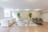 Located at 11 Margaret Ave in Kitchener, Ontario, this single-floor communal amenity space offers a generously sized, open-concept layout designed for gathering and relaxation. The space is divided into distinct functional zones that flow seamlessly into one another, all unified by warm light hardwood flooring and neutral grey-toned walls.

The main lounge area is a standout feature, highlighted by a striking stacked stone accent wall that adds texture and warmth to the room. Comfortable seating, including upholstered chairs and a sofa arranged around a coffee table on a geometric-patterned area rug, creates an inviting atmosphere. A large window and a glass door provide direct access to an outdoor patio area, filling the lounge with natural light and garden views.

Adjacent to the lounge, a dedicated dining area features multiple café-style round tables paired with modern blue upholstered chairs. Expansive windows line the wall, offering lush, tree-filled outdoor views and ensuring the space remains bright throughout the day.

The fully equipped kitchen includes ample warm-toned wooden cabinetry, dark granite countertops, stainless steel appliances, and large format tile flooring — providing everything needed for communal meal preparation. A window above the sink brings in additional natural light. The space also includes a smaller, flexible secondary room with hardwood flooring and its own seating area, suitable for private meetings or quiet use.