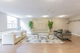 Located at 11 Margaret Ave in Kitchener, Ontario, this single-floor communal amenity space offers a generously sized, open-concept layout designed for gathering and relaxation. The space is divided into distinct functional zones that flow seamlessly into one another, all unified by warm light hardwood flooring and neutral grey-toned walls.

The main lounge area is a standout feature, highlighted by a striking stacked stone accent wall that adds texture and warmth to the room. Comfortable seating, including upholstered chairs and a sofa arranged around a coffee table on a geometric-patterned area rug, creates an inviting atmosphere. A large window and a glass door provide direct access to an outdoor patio area, filling the lounge with natural light and garden views.

Adjacent to the lounge, a dedicated dining area features multiple café-style round tables paired with modern blue upholstered chairs. Expansive windows line the wall, offering lush, tree-filled outdoor views and ensuring the space remains bright throughout the day.

The fully equipped kitchen includes ample warm-toned wooden cabinetry, dark granite countertops, stainless steel appliances, and large format tile flooring — providing everything needed for communal meal preparation. A window above the sink brings in additional natural light. The space also includes a smaller, flexible secondary room with hardwood flooring and its own seating area, suitable for private meetings or quiet use.