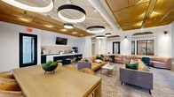 Located at 6220 Guilford Ave in Indianapolis, Indiana, this single-floor space offers a well-appointed, open-plan lounge and collaboration area designed with both comfort and function in mind. The interior design strikes a sophisticated balance between modern and warm aesthetics, highlighted by a striking gold-paneled ceiling feature that runs through the space and contrasts beautifully against the white exposed infrastructure above.

Oversized circular pendant lights hang at varying heights, creating a dramatic and layered lighting scheme throughout. The flooring transitions seamlessly between wood-look tile and patterned area rugs, defining distinct zones within the open layout.

A full kitchenette bar anchors one side of the space, featuring deep forest-green cabinetry with brushed gold hardware, a white quartz waterfall island with bar stools, subway tile backsplash, and a wall-mounted television—ideal for informal gatherings or presentations.

The lounge area accommodates multiple seating configurations, including caramel leather sofas, a curved blush pink banquette paired with a marble-top table, and velvet accent chairs in emerald green and slate blue. A long communal dining table with upholstered chairs sits near floor-to-ceiling windows fitted with roller shades, allowing diffused natural light to fill the space. The overall ambiance is warm, inviting, and professionally curated.