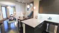 This thoughtfully designed single-floor space showcases an open-concept layout that seamlessly connects the kitchen, dining, and living areas into one cohesive and inviting environment. The modern kitchen is a standout feature, boasting a generous granite-topped island with sleek dark cabinetry and brushed metal hardware. Pendant lights hang above the island, casting a warm glow that enhances the space's contemporary aesthetic. The kitchen flows naturally into the dining area, where an industrial-style table and metal chairs create a chic yet functional gathering spot. Bold, abstract artwork adorns the walls, adding a dynamic visual element and a touch of artistic character to the interior. Sheer floor-to-ceiling curtains filter natural light softly throughout the space, contributing to an airy and open ambiance. Beyond the dining area, the living room offers a comfortable retreat, further enriched by the continuity of the polished dark hardwood flooring that runs throughout. The overall design balances modern sophistication with warm, livable comfort, making this space as practical as it is visually appealing.