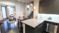 This thoughtfully designed single-floor space showcases an open-concept layout that seamlessly connects the kitchen, dining, and living areas into one cohesive and inviting environment. The modern kitchen is a standout feature, boasting a generous granite-topped island with sleek dark cabinetry and brushed metal hardware. Pendant lights hang above the island, casting a warm glow that enhances the space's contemporary aesthetic. The kitchen flows naturally into the dining area, where an industrial-style table and metal chairs create a chic yet functional gathering spot. Bold, abstract artwork adorns the walls, adding a dynamic visual element and a touch of artistic character to the interior. Sheer floor-to-ceiling curtains filter natural light softly throughout the space, contributing to an airy and open ambiance. Beyond the dining area, the living room offers a comfortable retreat, further enriched by the continuity of the polished dark hardwood flooring that runs throughout. The overall design balances modern sophistication with warm, livable comfort, making this space as practical as it is visually appealing.