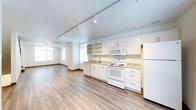 This multi-floor space spans three levels, offering a versatile and functional layout designed for comfortable living. The main living and kitchen area showcases an open-concept design, where the kitchen flows seamlessly into the living space. The kitchen is equipped with white cabinetry, open shelving, granite-style countertops, and full-size white appliances including a range, microwave, and refrigerator. A deep sink with a single-handle faucet is positioned beneath a window, allowing natural light to fill the workspace. The flooring throughout features wood-style planks in a warm, neutral tone that adds warmth and continuity across the space. Track lighting runs along the ceiling, providing adjustable, directed illumination to complement the natural light pouring in through multiple windows fitted with horizontal blinds. A ceiling fan is visible in the adjoining living area, adding both comfort and a functional design element. The overall aesthetic is clean, modern, and move-in ready, with a bright, airy ambiance enhanced by white walls and trim. Spread across three floors, this space provides ample room for a variety of living arrangements and lifestyle needs.