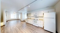 This multi-floor space spans three levels, offering a versatile and functional layout designed for comfortable living. The main living and kitchen area showcases an open-concept design, where the kitchen flows seamlessly into the living space. The kitchen is equipped with white cabinetry, open shelving, granite-style countertops, and full-size white appliances including a range, microwave, and refrigerator. A deep sink with a single-handle faucet is positioned beneath a window, allowing natural light to fill the workspace. The flooring throughout features wood-style planks in a warm, neutral tone that adds warmth and continuity across the space. Track lighting runs along the ceiling, providing adjustable, directed illumination to complement the natural light pouring in through multiple windows fitted with horizontal blinds. A ceiling fan is visible in the adjoining living area, adding both comfort and a functional design element. The overall aesthetic is clean, modern, and move-in ready, with a bright, airy ambiance enhanced by white walls and trim. Spread across three floors, this space provides ample room for a variety of living arrangements and lifestyle needs.