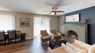 This single-floor space is thoughtfully laid out across several distinct areas, each with its own character and function. The living room serves as the heart of the home, anchored by a striking stone fireplace with a built-in shelf and textured accent wall. Wood-style flooring runs throughout, complementing the warm, neutral tones of the walls. A ceiling fan overhead adds comfort, while a sliding glass door fitted with vertical blinds opens to a view of an outdoor pool, filling the room with natural light and a refreshing outdoor connection.

The kitchen is well-appointed with light wood cabinetry offering generous storage, dark countertops, and a full suite of white appliances including a refrigerator, electric range, and built-in microwave. Wood-style flooring continues here, and a window with vertical blinds provides additional natural light. A dining area adjacent to the kitchen features patterned carpet, a ceiling fan, and a window unit for climate control, providing a comfortable and functional space for everyday dining.

Throughout the unit, textured ceilings, neutral wall tones, and consistent design choices create a cohesive and practical living environment suitable for comfortable, everyday living.