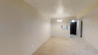 This 1-bedroom, 1-bathroom unit at Pineridge is thoughtfully laid out on a single floor, making it easy to navigate and functional for everyday living. The main living area features neutral beige carpeting and cream-colored walls that create a bright, clean backdrop. A small chandelier serves as a focal point overhead, adding a subtle decorative touch to the open space. A pass-through window connects the living area to the kitchen, improving the flow between spaces and allowing light to travel through the unit. The kitchen is visible through the pass-through and features dark cabinetry, offering a contrast to the lighter tones of the main living space. A ceiling fan is installed in the adjacent area, providing additional air circulation. The overall color palette throughout the unit remains consistent — light, warm neutrals that lend the space a calm and inviting atmosphere. The layout is compact yet efficient, designed to maximize usability within its footprint. This unit is well-suited for an individual or couple seeking a manageable, low-maintenance living space with a straightforward, practical design.