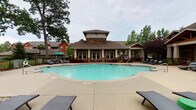 Located at Legacy Oaks Place in Asheville, North Carolina, this outdoor amenity area offers a well-appointed resort-style retreat designed for relaxation and socializing. The centerpiece is a generously sized, freeform swimming pool with clear turquoise water, surrounded by an expansive concrete pool deck furnished with wicker chaise lounges and shaded by mature oak and ornamental trees. The adjacent clubhouse building features craftsman-style architecture with stone accents, a raised cupola, large windows, and covered porticos that extend the indoor-outdoor living experience. A standout feature of the space is the covered pavilion, constructed with heavy timber framing and a vaulted wood ceiling, anchored by a striking floor-to-ceiling stacked stone fireplace equipped with a mounted flat-screen television above the hearth. The pavilion also includes a stone-clad outdoor bar and kitchen area, complemented by comfortable seating arrangements with navy blue cushions. Thoughtfully designed landscaping with ornamental shrubs, flowering plants, and mulched garden beds frames the entire area, while wrought iron fencing provides definition and security. Stone pillar accents and vintage-style pole lighting add charm and cohesion to this well-maintained outdoor gathering space.