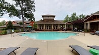 Located at Legacy Oaks Place in Asheville, North Carolina, this outdoor amenity area offers a well-appointed resort-style retreat designed for relaxation and socializing. The centerpiece is a generously sized, freeform swimming pool with clear turquoise water, surrounded by an expansive concrete pool deck furnished with wicker chaise lounges and shaded by mature oak and ornamental trees. The adjacent clubhouse building features craftsman-style architecture with stone accents, a raised cupola, large windows, and covered porticos that extend the indoor-outdoor living experience. A standout feature of the space is the covered pavilion, constructed with heavy timber framing and a vaulted wood ceiling, anchored by a striking floor-to-ceiling stacked stone fireplace equipped with a mounted flat-screen television above the hearth. The pavilion also includes a stone-clad outdoor bar and kitchen area, complemented by comfortable seating arrangements with navy blue cushions. Thoughtfully designed landscaping with ornamental shrubs, flowering plants, and mulched garden beds frames the entire area, while wrought iron fencing provides definition and security. Stone pillar accents and vintage-style pole lighting add charm and cohesion to this well-maintained outdoor gathering space.