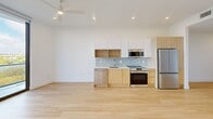 This thoughtfully designed 1-bedroom, 2-bathroom residence offers a clean, contemporary aesthetic across a single floor. Light hardwood flooring runs throughout, creating a cohesive and airy feel. The open-concept kitchen and living area serves as the heart of the home, featuring a well-appointed kitchen with a two-tone cabinetry system — white upper cabinets paired with warm wood lower cabinets — complemented by a soft blue tile backsplash, stainless steel appliances including a refrigerator, range, microwave, and dishwasher, and recessed lighting above. A ceiling fan adds to the comfort of the living space. Floor-to-ceiling windows and a glass-railed balcony access in the living area flood the space with natural light and offer expansive outdoor views of the surrounding landscape. The bedroom continues the minimalist design with large-format windows that frame sweeping views. The primary bathroom is a standout feature, boasting a wide double-sink vanity with warm wood cabinetry, a white quartz countertop, and a large LED-backlit mirror that adds a modern, luminous touch. A hallway connects the rooms efficiently, with louvered closet doors providing both function and style.