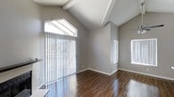 This single-floor space makes a strong first impression with its dramatic vaulted ceilings accented by exposed decorative beams, creating a sense of openness and height that defines the overall atmosphere. The main living area is bathed in natural light, courtesy of a large sliding glass door fitted with vertical blinds and flanked by transom windows that draw the eye upward toward the soaring roofline. Additional windows along the adjacent wall further enhance the brightness and airy feel of the space. Smooth, neutral gray walls provide a clean, contemporary backdrop that complements the warm tones of the rich, dark hardwood flooring, which runs throughout and adds a sense of elegance and warmth. White baseboards and trim offer a crisp contrast, giving the interior a polished, well-finished look. The open layout promotes a flexible use of the space, while the high ceiling contributes to a feeling of expansiveness well beyond what the footprint alone might suggest. The combination of modern finishes, natural light, and architectural detailing makes this a welcoming and visually compelling space suited to a variety of uses.